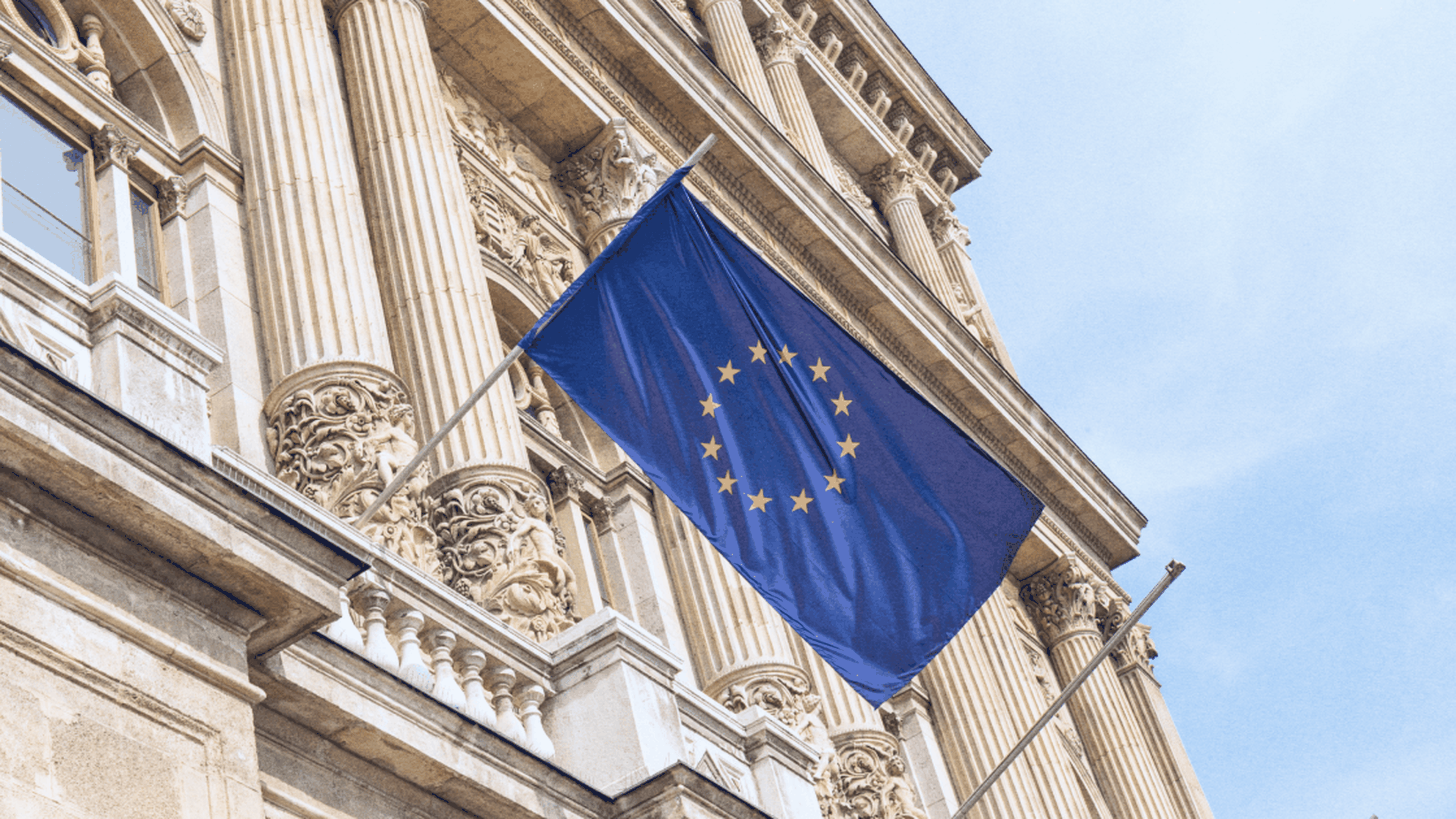 European Union flag with gold stars on a blue background waving outside a classical building with carved columns.