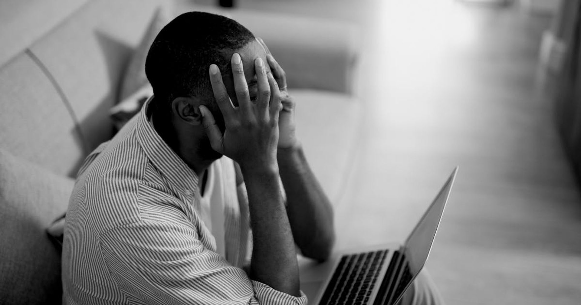 A black man frustratingly holding his head in his hands while looking at a laptop