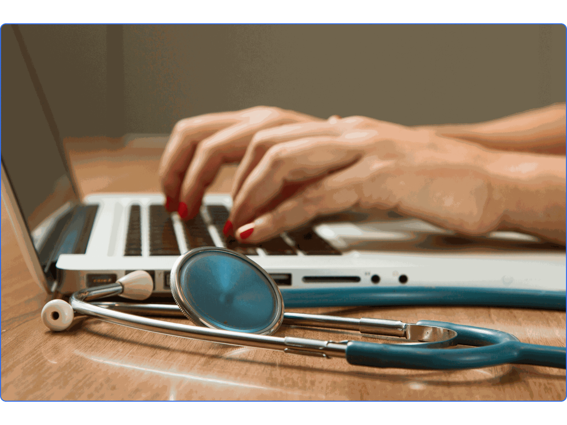 Hands typing on a laptop keyboard with a stethoscope nearby on a wooden table.