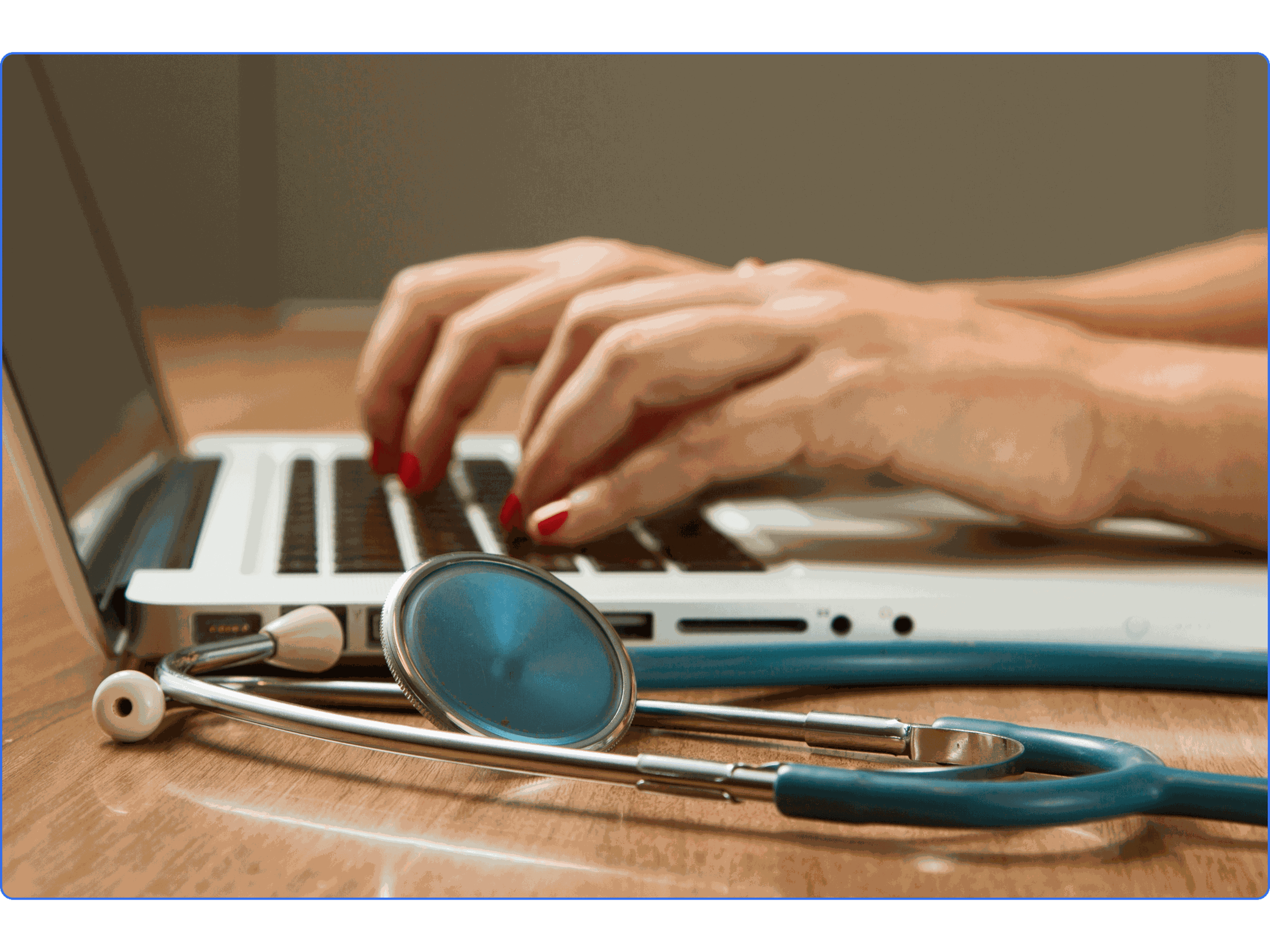 Hands typing on a laptop keyboard with a stethoscope nearby on a wooden table.