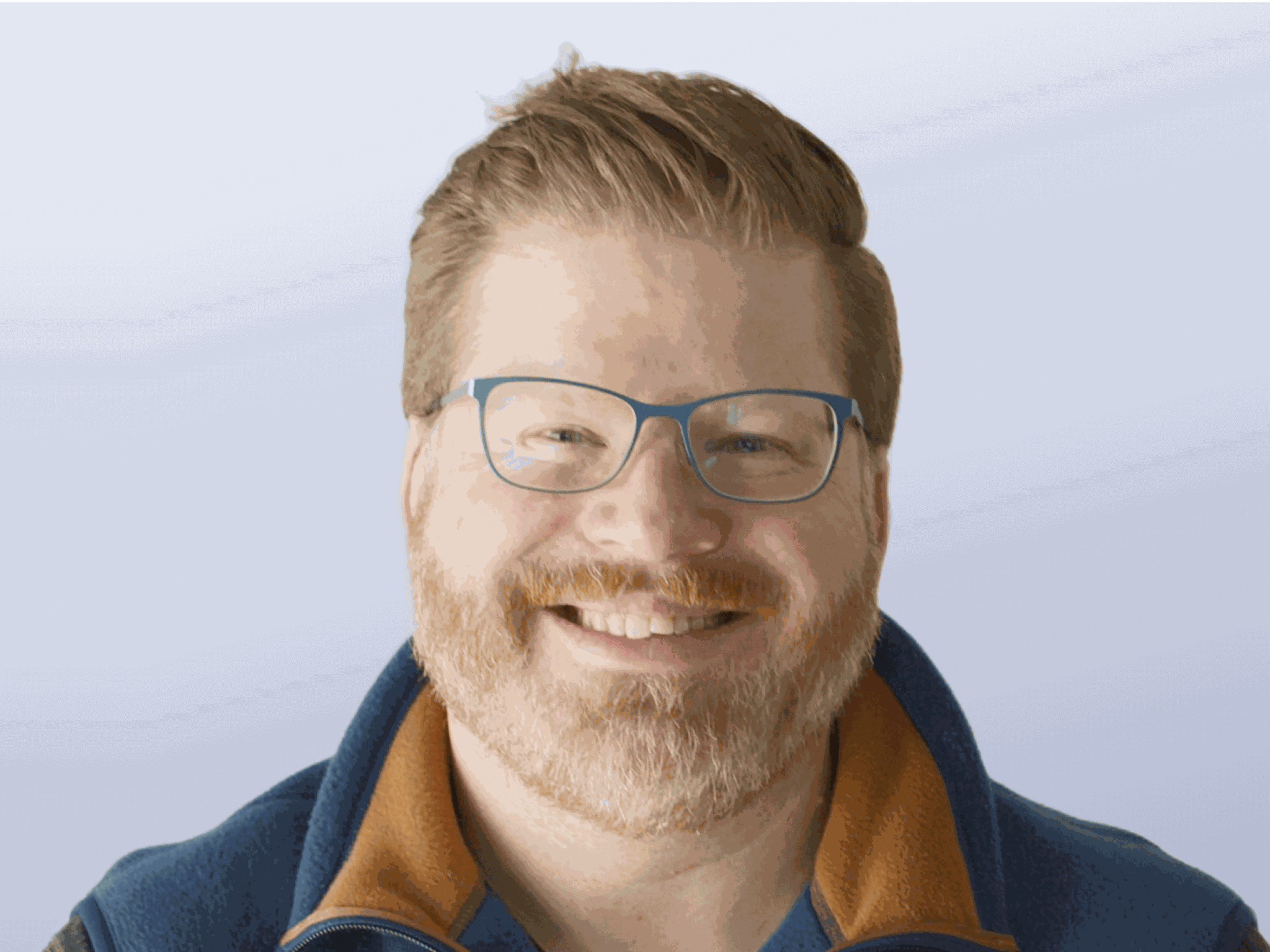 A headshot of John Postlethwait. A caucasian male with medium red hair and facial hair wearing a blue and orange zip-up jacket.
