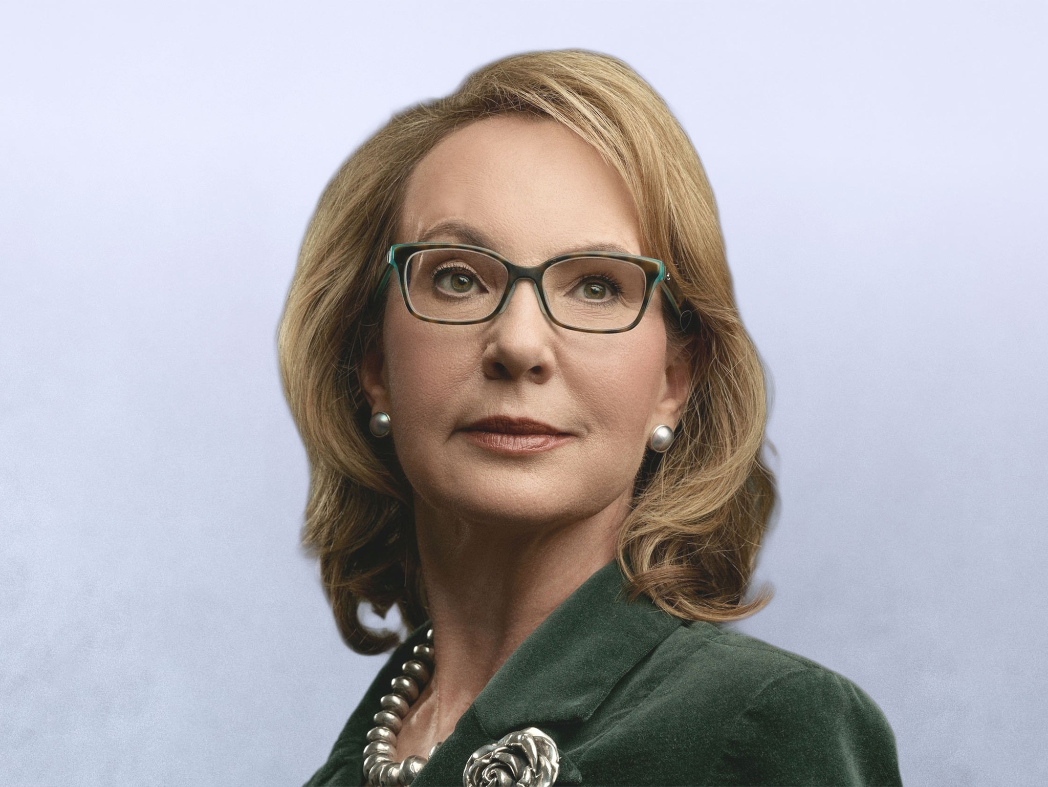 A headshot of Gabby Giffords. A caucasian woman with shoulder length blond hair, wearing a green blazer and glasses.
