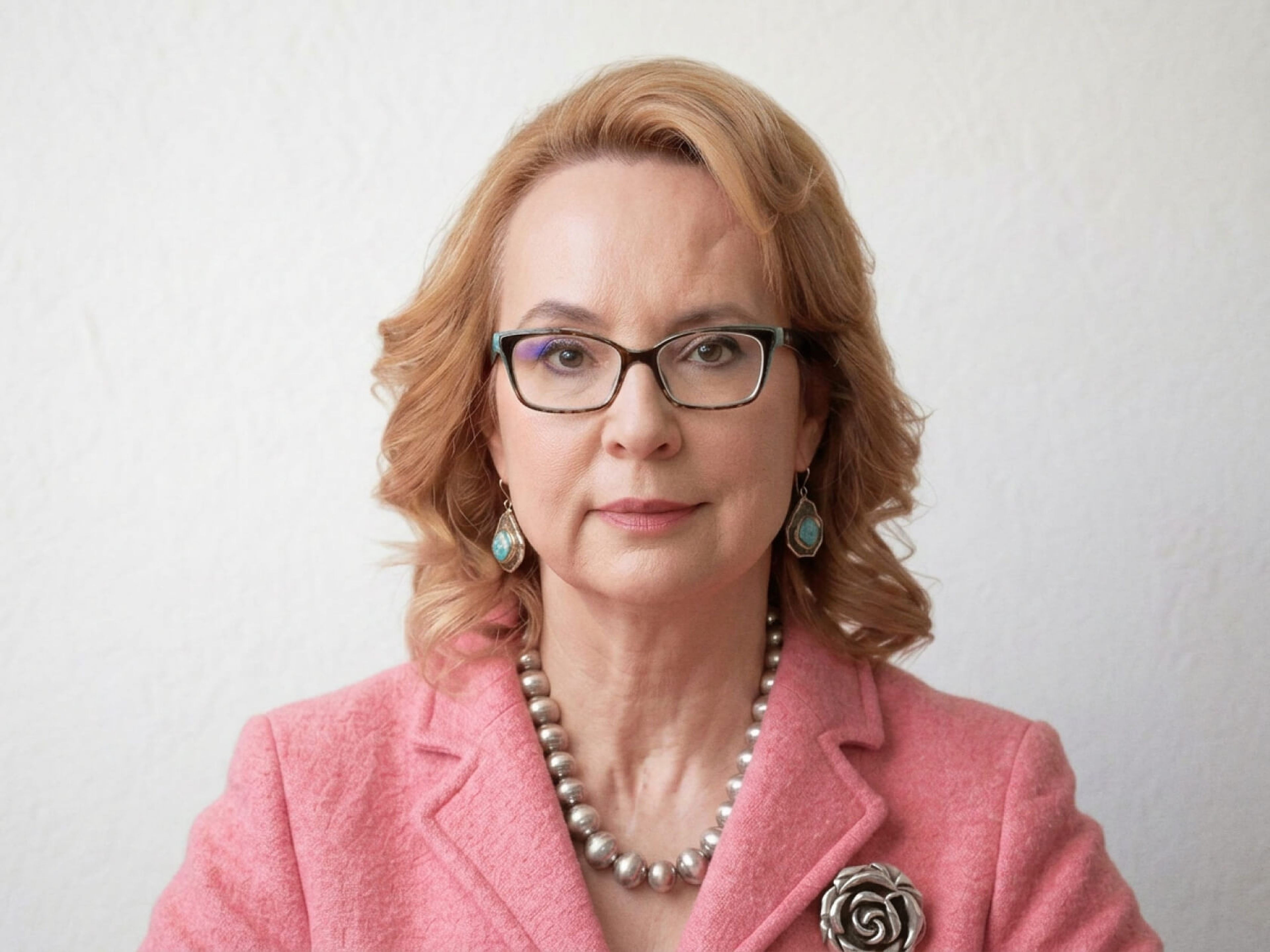 A headshot of Gabby Giffords. A caucasian woman with shoulder length blond hair, wearing a green blazer and glasses.