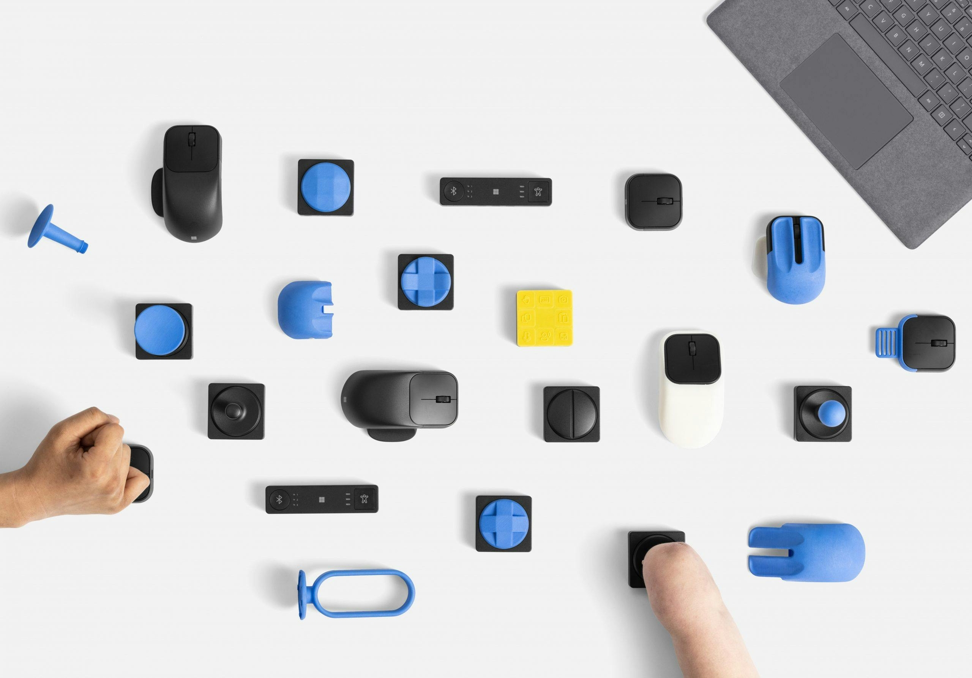 A top-down view of different Microsoft inclusive tech accessories laid on a table, with two people using the different peripherals.