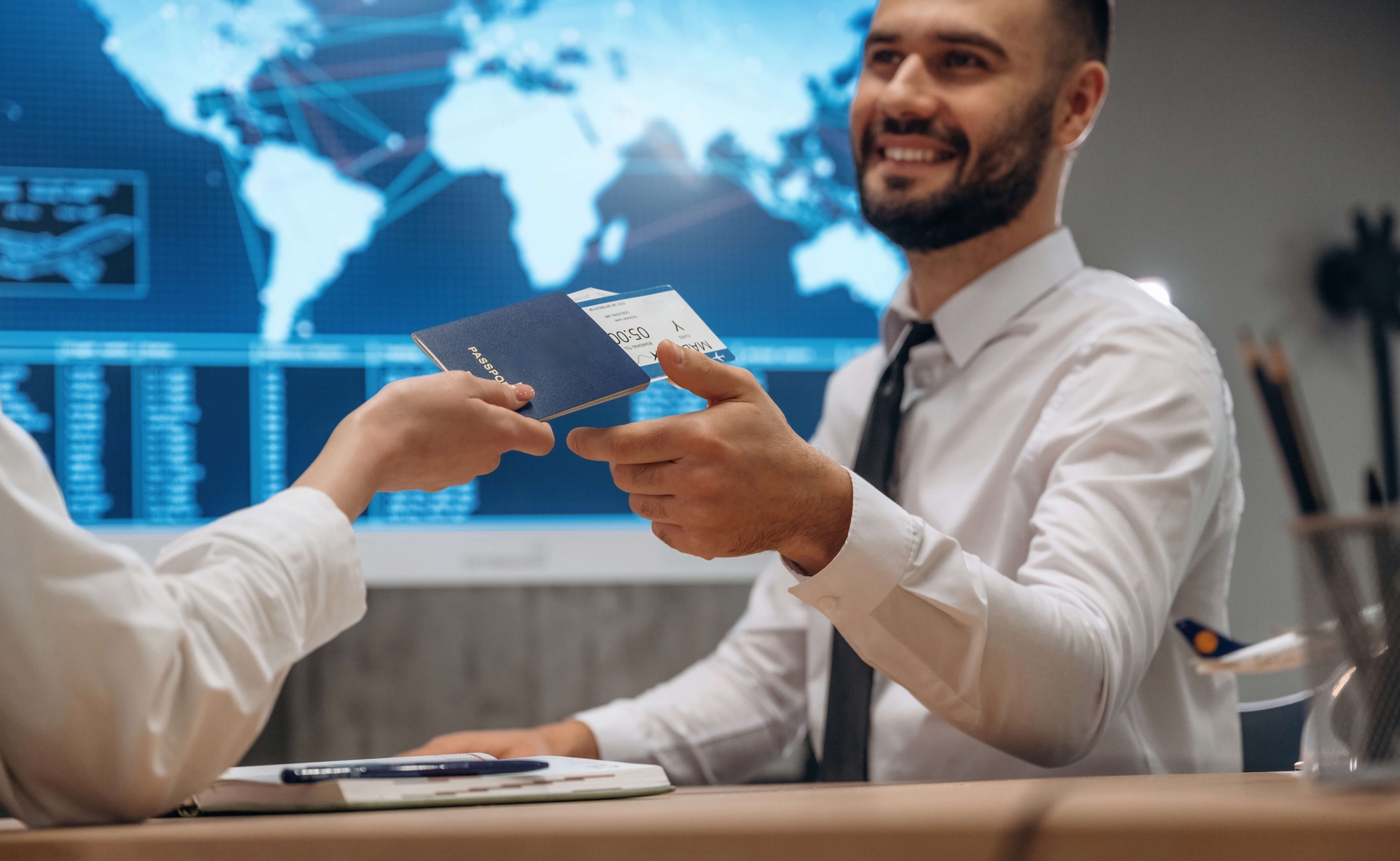 A smiling man hands over a passport and boarding pass at a travel desk, with a world map and flight data in the background.