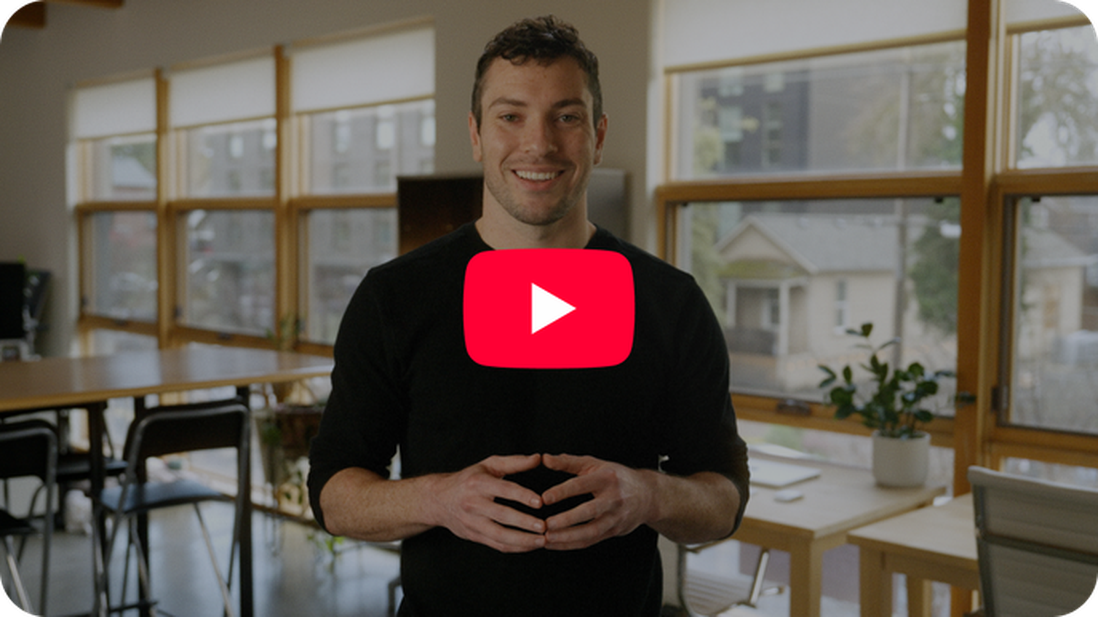 Man in a black shirt smiling and standing in a modern room with large windows, tables, and a plant. A YouTube play button overlays the image.
