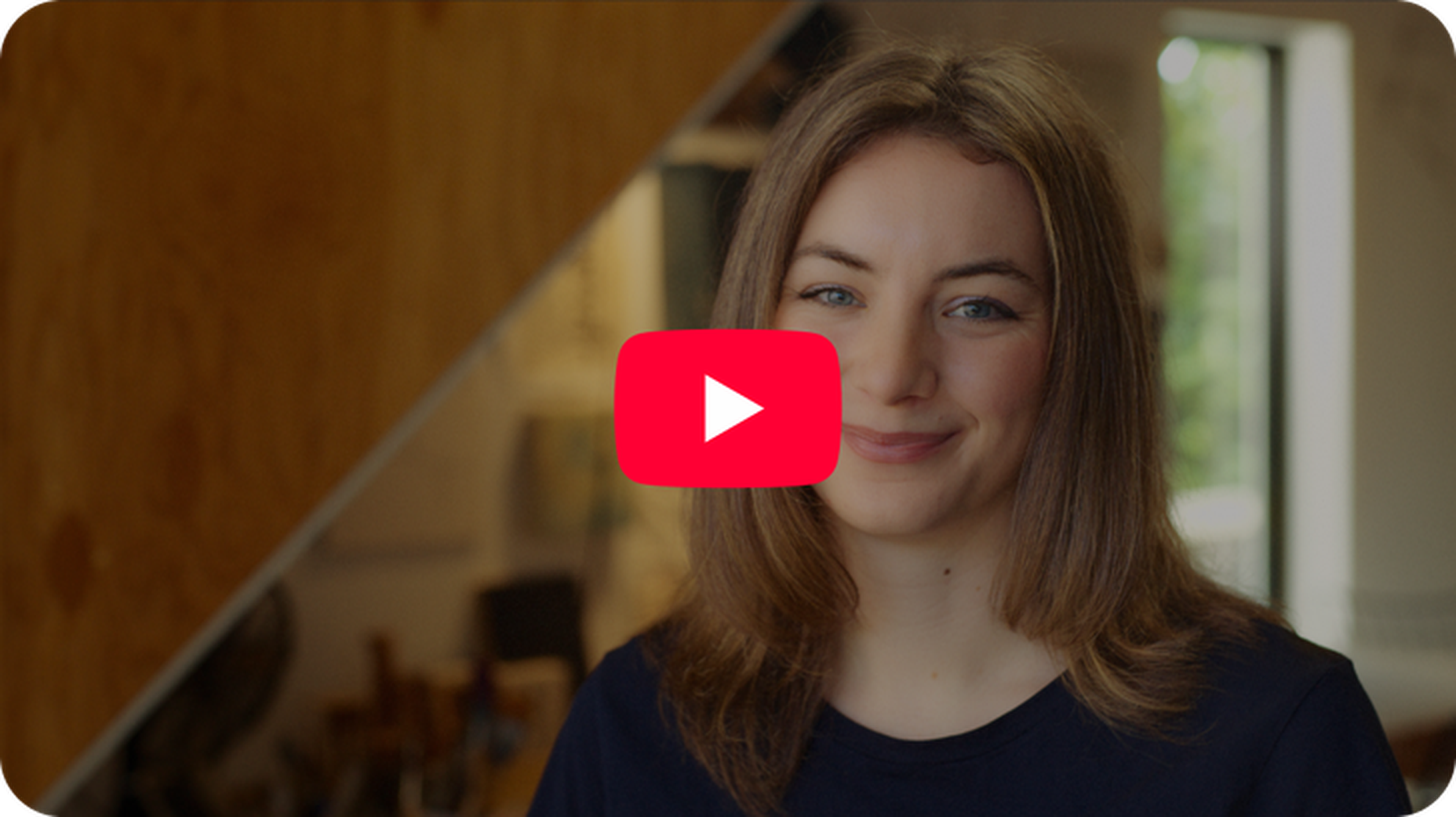 Woman with long brown hair smiling indoors, with blurred background and red play button overlay.